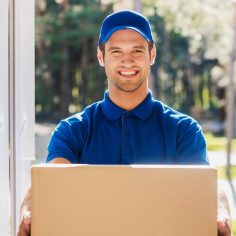 Take your package! Happy young delivery man stretching out a cardboard box while standing at the entrance of apartment